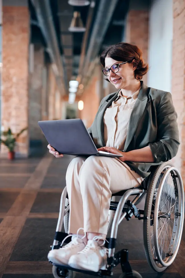 Eine lächelnde Frau im Rollstuhl sitzt in einem modernen Bürogebäude und arbeitet an ihrem Laptop.