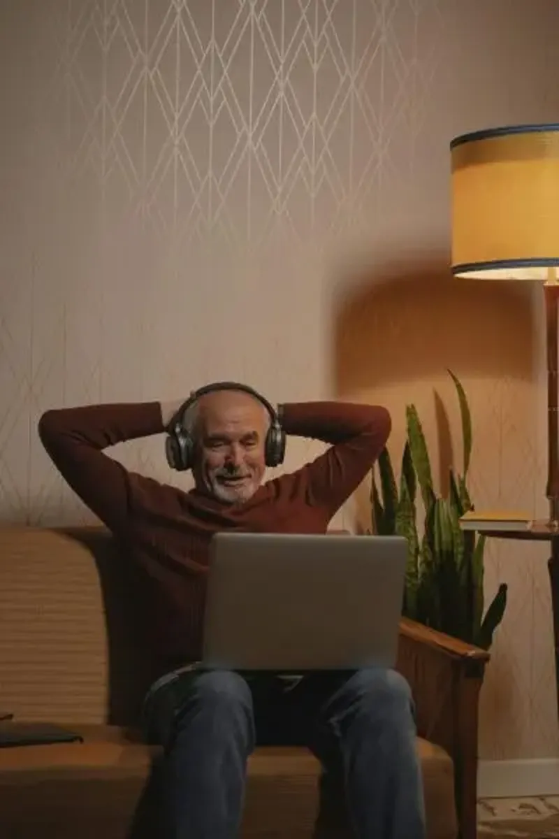 Älterer Mann mit Kopfhörern lehnt sich entspannt auf einem Sofa zurück, während er auf seinen Laptop schaut. Eine warme Lichtquelle von einer Stehlampe sorgt für eine gemütliche Atmosphäre.