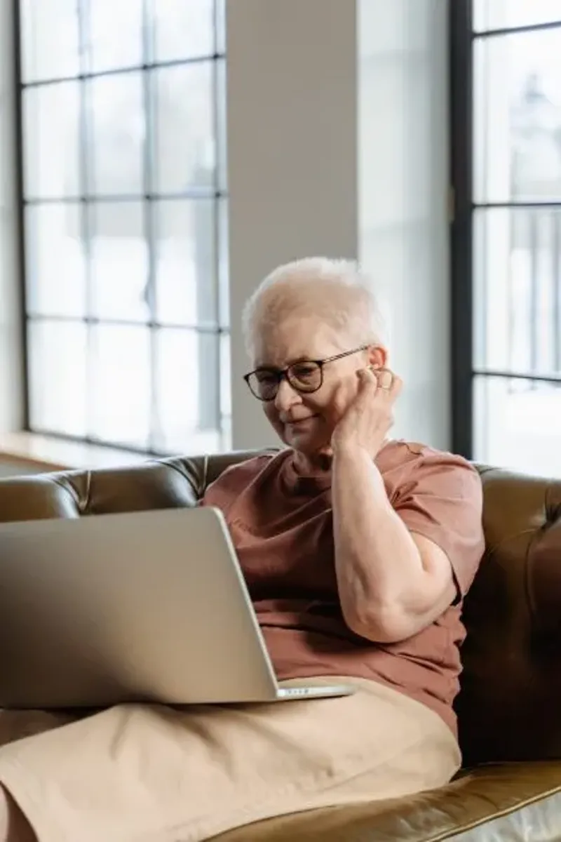 Eine ältere Frau mit kurzen weißen Haaren und Brille sitzt entspannt auf einem Ledersofa und nutzt ihren Laptop. Sie trägt ein beigefarbenes Kleid und ein braunes T-Shirt. Während des Gesprächs fasst sie sich ans Ohr, als ob sie ein Hörgerät oder Kopfhörer anpasst. Große Fenster im Hintergrund lassen viel Tageslicht in den Raum.
