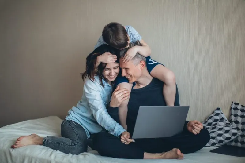 Lächelnde Familie sitzt gemeinsam auf dem Bett, während sie auf einen Laptop schauen. Das Kind sitzt auf den Schultern des Vaters und umfasst liebevoll die Köpfe der Eltern.