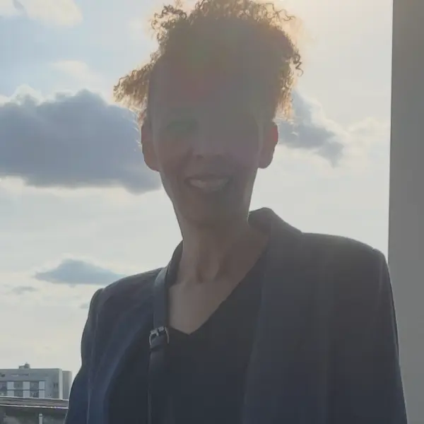 Bettina Anderson im blauen Blazer und schwarzem Top; lockiges Haar, draußen vor hellem, wolkigem Himmel.